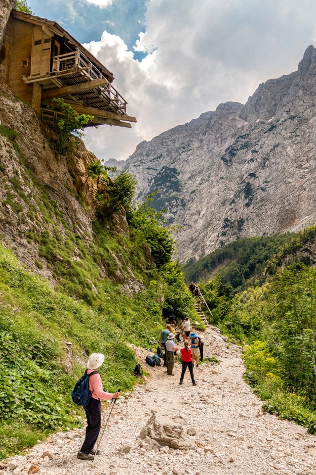 Hikers embark on a wild and rugged adventure on a rocky mountain trail surrounded by green vegetation. A wooden cabin is built into the cliffside above them. Snow-capped peaks and a partly cloudy sky grace the background, capturing the untamed beauty of Slovenia.