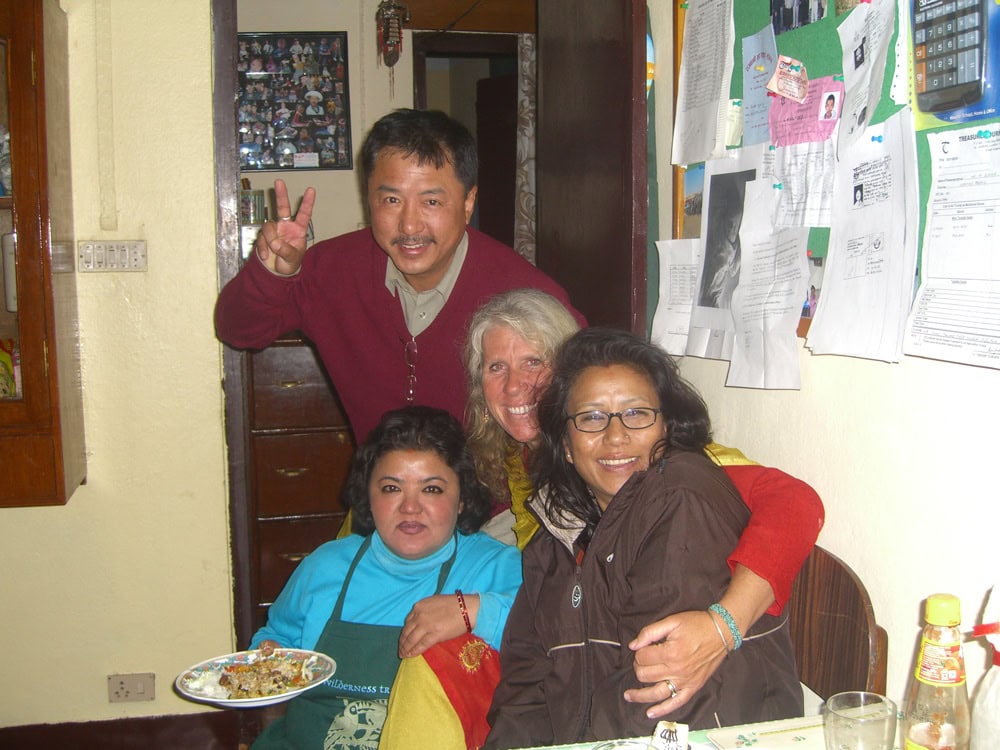 Four people are gathered indoors; three are seated, and one stands behind, making a peace sign with his fingers. Various papers, photos, and notes cover the wall in the background, as if part of a family photobook documenting their recent trip to Nepal.