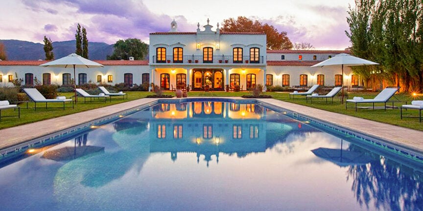 A large white villa with arched windows and doors, surrounded by a green lawn, sun loungers, and white umbrellas. The rectangular swimming pool in the foreground reflects the building, blending elegance and the spirit of adventure typically found in wine-growing regions.