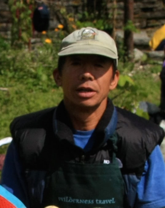 A person wearing a green cap and a black vest poses outdoors with lush greenery in the background, perfect for a family photobook.