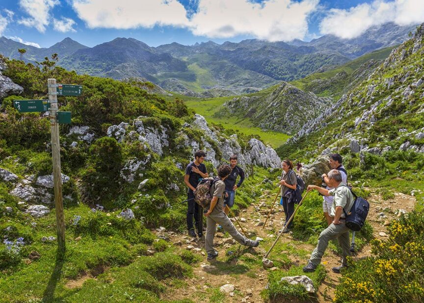 A group of hikers stands at a trail intersection surrounded by green mountains under a partly cloudy European sky. They appear to be discussing or taking a break. A signpost with trail directions is visible, guiding more enthusiastic crowds through the scenic trails.