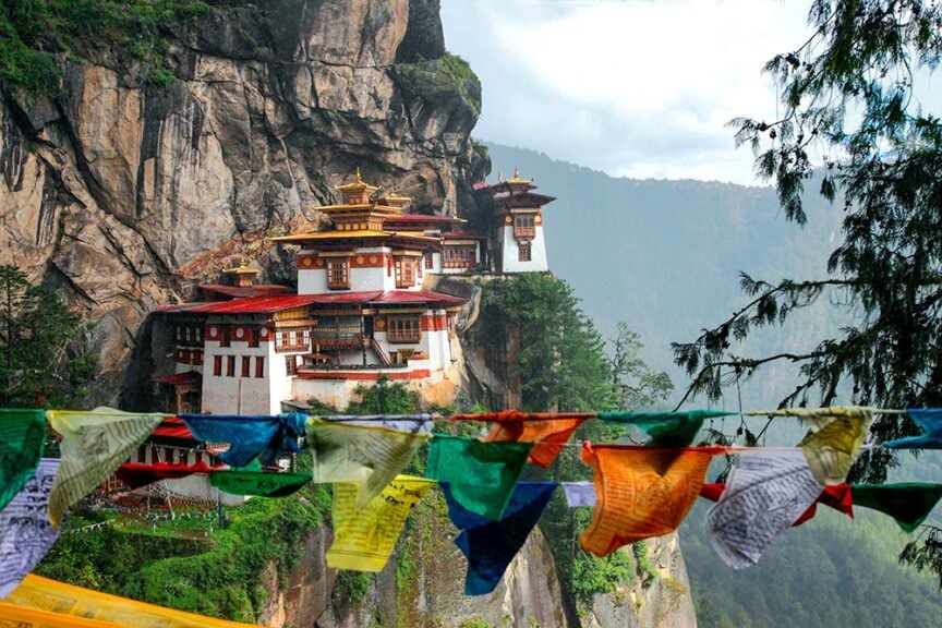 The image shows the Tiger's Nest Monastery, a cliffside Buddhist monastery in Bhutan, with colorful prayer flags in the foreground and mountains in the background, capturing an adventurous hiking trip that offers breathtaking views and spiritual tranquility.