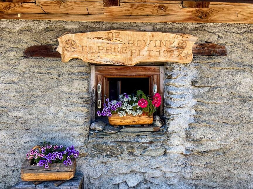 A small stone window adorned with vibrant flowers in wooden planters, reminiscent of scenes from Tour du Mont Blanc. Above the window is a wooden sign with inscriptions. Another planter with purple flowers is placed on a ledge below, perfect for capturing trail photos.