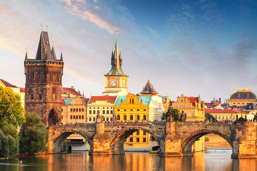 A historic stone bridge spans a river, connecting buildings with varied architectural styles under a partly cloudy sky, often attracting crowds of tourists exploring Europe.