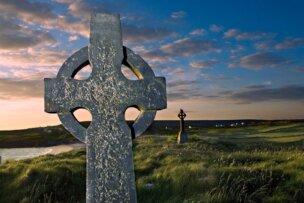A weathered stone Celtic cross stands in a grassy field during sunset in Inishmore, Ireland, with another cross visible in the distance.