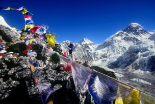 Colorful prayer flags flutter in the wind against a backdrop of snow-covered mountains and a bright blue sky in Nepal. A climber, embodying the spirit of exploration, stands among the rocks in the background.