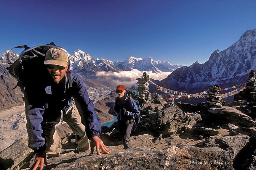 Two hikers wearing backpacks trek up a rocky mountain path in Nepal, with the Himalayan range and prayer flags in the background under a clear blue sky, embodying the spirit of exploration.