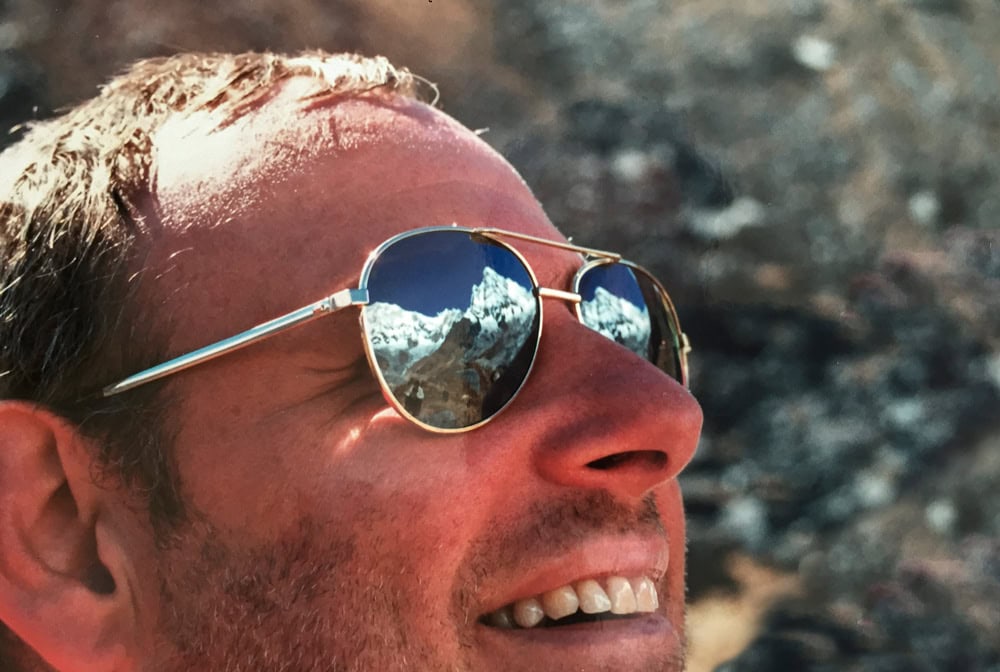 A person wearing reflective sunglasses smiles with a mountainous landscape of Nepal mirrored in the lenses, capturing a perfect moment for the family photobook.