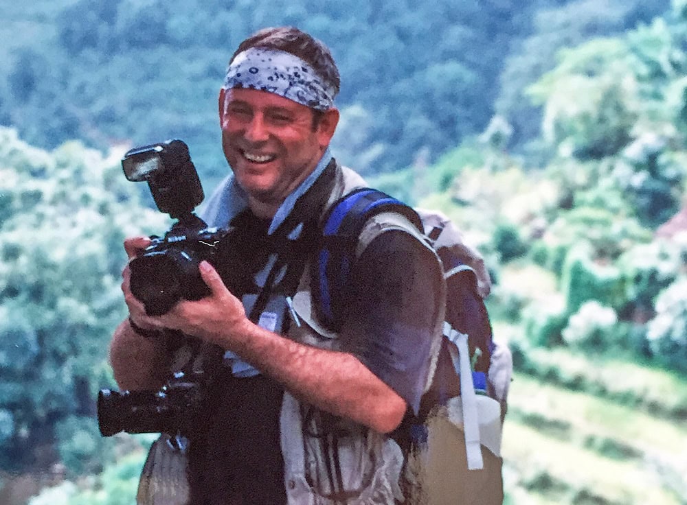 A person wearing a bandana and a backpack is smiling while holding a camera with a flash attachment, perhaps capturing moments for a family photobook. The background features a blurred green, forested landscape, reminiscent of Nepal's breathtaking scenery.