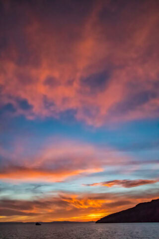 A vibrant sunset with a mix of orange, pink, and purple clouds over the calm Sea of Cortez, featuring a silhouetted boat and the distant coastline of Baja by Boat.