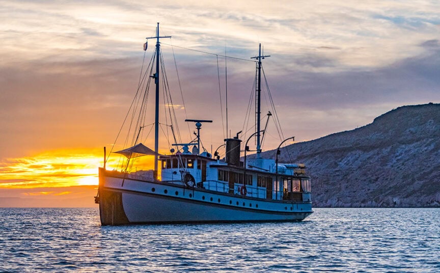 A large boat floats on calm water at sunset, with a mountainous shoreline in the background. The sky is partly cloudy, and the sun is low on the horizon, creating a serene scene perfect for boat exploring in Baja's stunning Sea of Cortez.