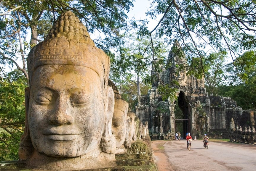 Stone faces carved into statues line a path leading to an ancient temple entrance, evoking the mystique of Urban Antiquity; two cyclists ride toward the temple under tree branches.