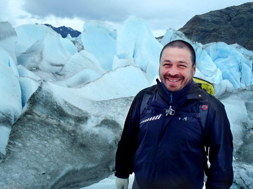 Carlos Leon, in a black jacket, stands smiling in front of blue and white glacier formations under a cloudy sky for his personal profile.