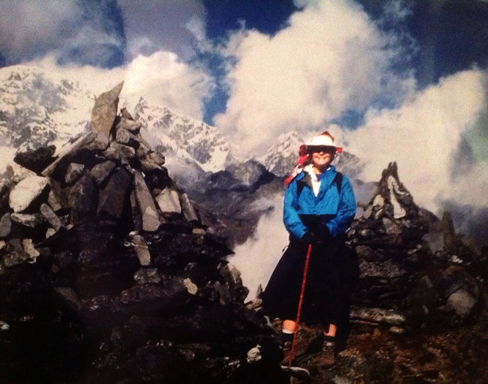 A person in outdoor gear stands with a walking stick between two rock formations, with snow-capped mountains and clouds in the background, capturing a moment perfect for a family photobook from a trek in Nepal.