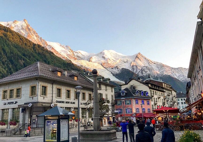 A town square with people and buildings, set against a background of snow-capped mountains at sunset in Chamonix-Mont-Blanc, France. The mighty peaks invite adventurers planning their next hike or gearing up for the renowned Tour du Mont Blanc trail.