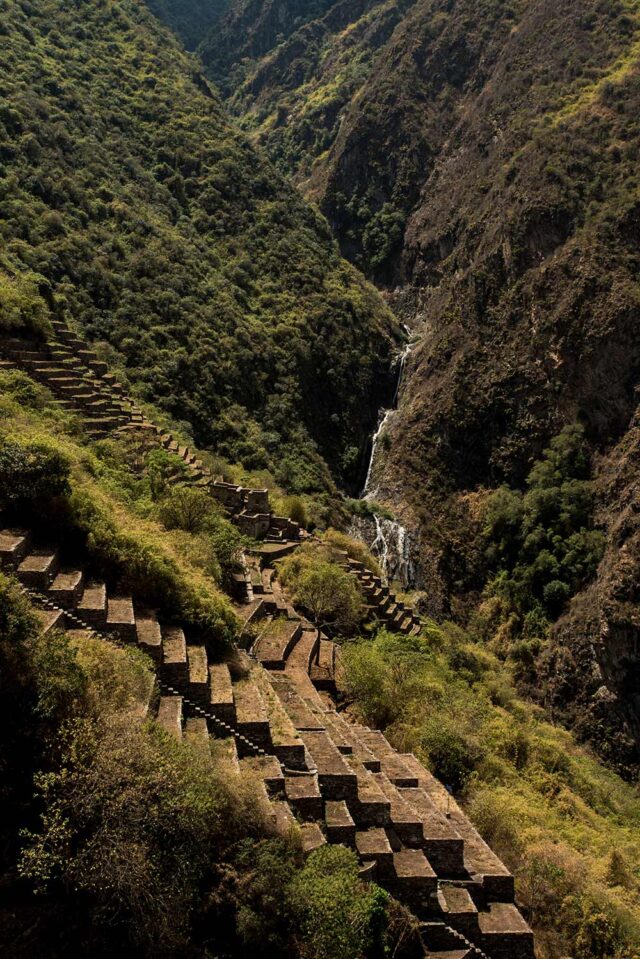 Terraced stone steps lead up a green, hilly landscape with a waterfall in the background, leaving visitors in awe of the beauty reminiscent of Choquequirao.