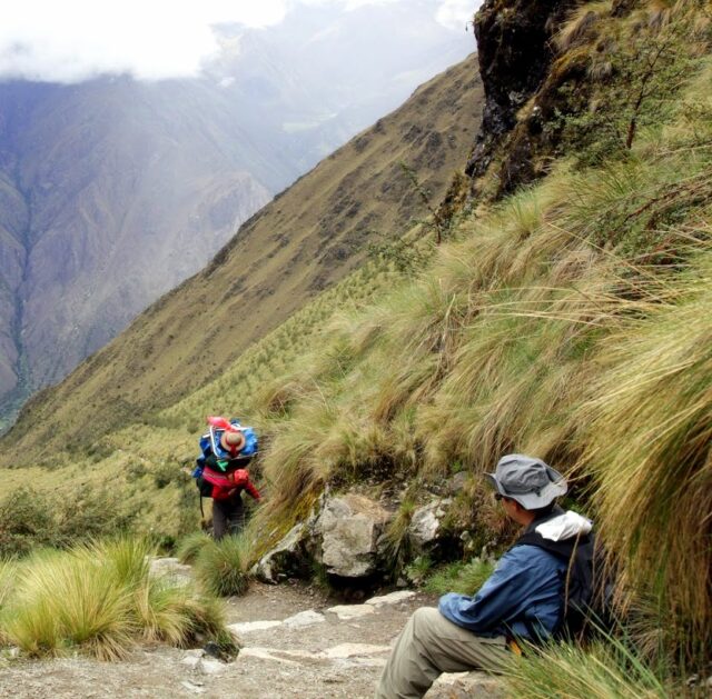 Two hikers traverse a steep mountain trail. One hiker, following the leader with a baby carrier on their back, ascends steadily, while the other sits on a grassy slope, taking a break. Clouds and mountains dominate the background.