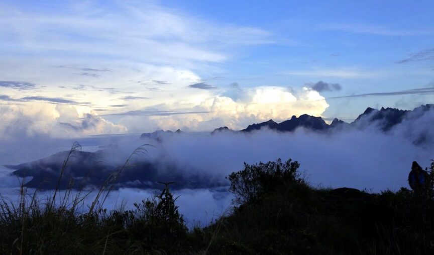 Mountain peaks rise majestically under a blue sky with scattered clouds. In the foreground, lush vegetation follows the contours of the landscape, leading the eye to nature's grandeur.