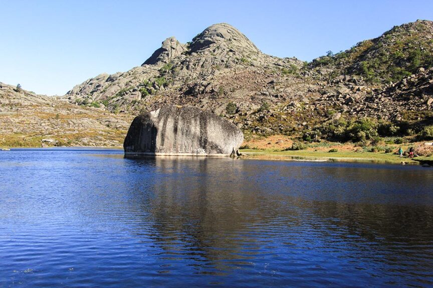 A large rock formation stands in the middle of a calm lake surrounded by mountainous terrain and clear skies, embodying the simple pleasures of rural Portugal.