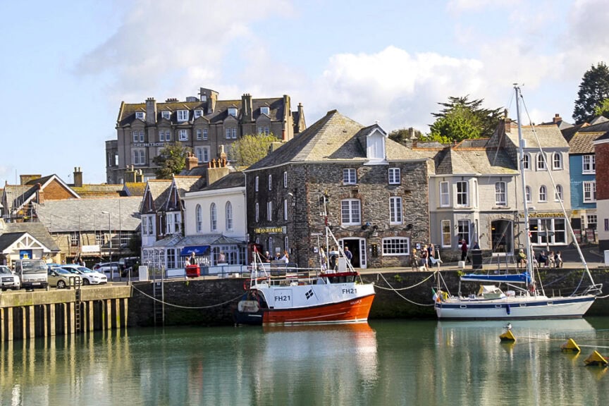 A scenic harbor in England features moored boats, a row of charming stone and brick buildings, and a large building on a hill in the background under a partly cloudy sky. Nearby trails wind through quaint villages, adding to the picturesque setting.