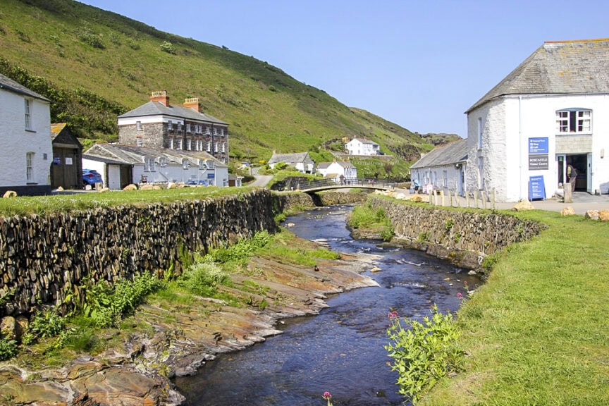 A narrow stream runs through a charming village in England, with grassy banks, stone houses, and a stone wall on one side, set against a backdrop of green hills and scenic trails under a clear blue sky.