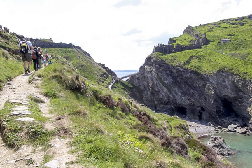 A group of hikers walks along England's Trails on a grassy cliffside with a rocky landscape, overlooking a body of water and a distant bridge.