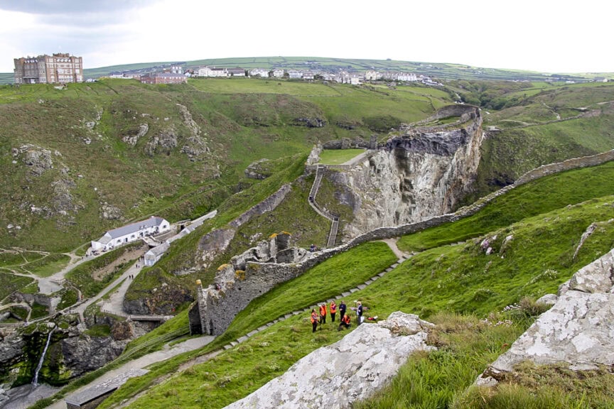Castle ruins on a cliffside with green hills and a building in the background. A group of people in bright clothing are gathered at the bottom of the hill. Trails and walls weave through the landscape, reminiscent of charming villages found throughout England.