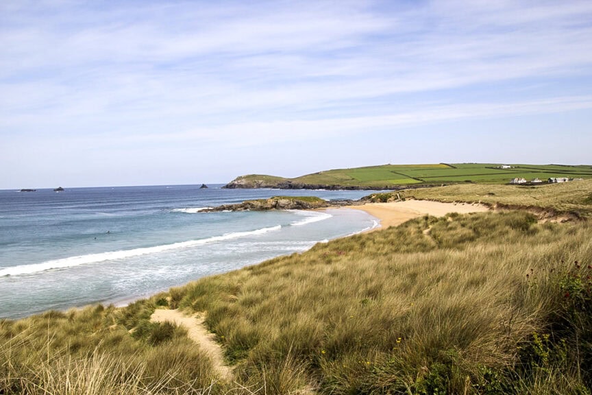 Coastal landscape featuring a sandy beach, rolling waves, and grassy dunes under a blue sky. Prominent green fields and distant rocky outcrops complete the scene, typical of charming villages in England with winding trails inviting exploration.
