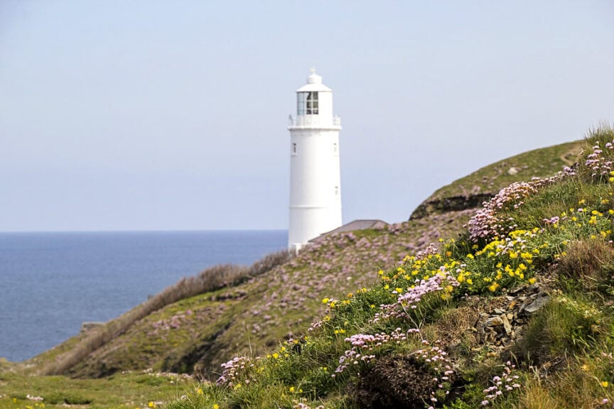 A white lighthouse stands on a grassy hill in one of England's charming villages, overlooking the sea with colorful wildflowers in the foreground and a clear sky above, perfect for exploring nearby trails.