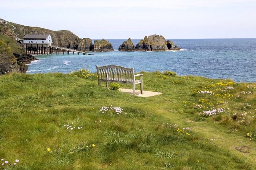 A wooden bench overlooks a cliffside view of the ocean and rocky islets, with a building on stilts extending into the sea on the left. The scene includes grassy terrain with scattered flowers, reminiscent of England's charming villages and scenic trails.