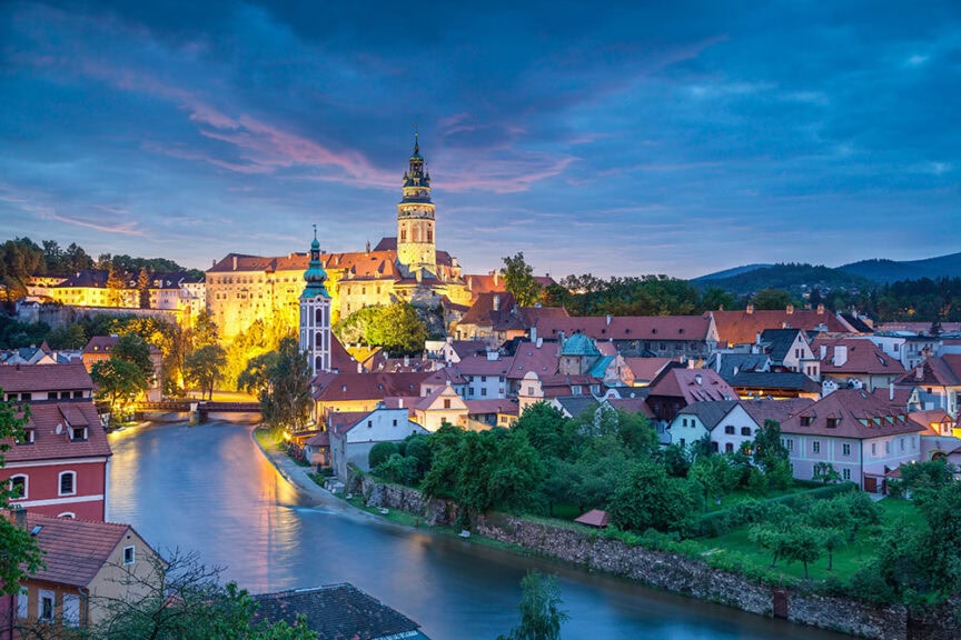 A picturesque view of a European town at dusk, with a river flowing through, historic buildings, and a tall church tower illuminated against a blue sky. Nestled in the background are ancient castles that add to the enchanting atmosphere.