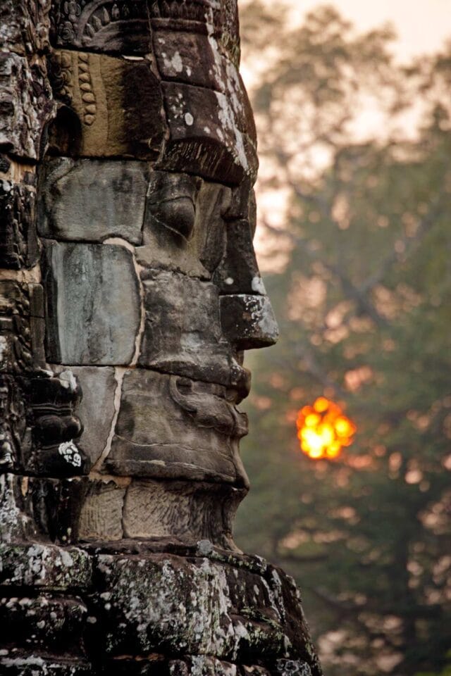Close-up of an ancient stone face sculpture with a serene expression at sunrise, captured at Angkor Wat, with the sun partially visible through trees in the background.