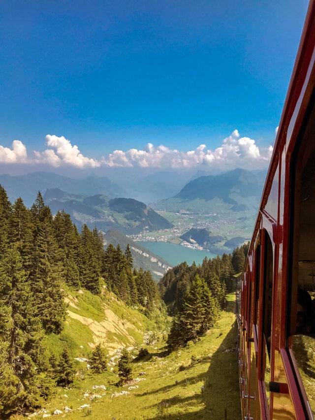 A red train travels on a mountain in Switzerland, passing through a forest with a scenic view of a lake and distant peaks under a blue sky with scattered clouds. This picturesque route also intersects with the Via Alpina, inviting hiking enthusiasts to explore the trails.