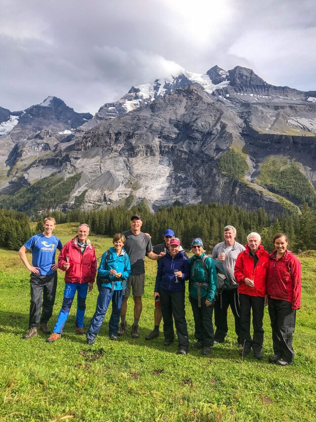 A group of nine people in outdoor clothing stand on a grassy area with a mountainous, partially snowy background, likely enjoying a day of hiking along the scenic trails of Via Alpina in Switzerland.