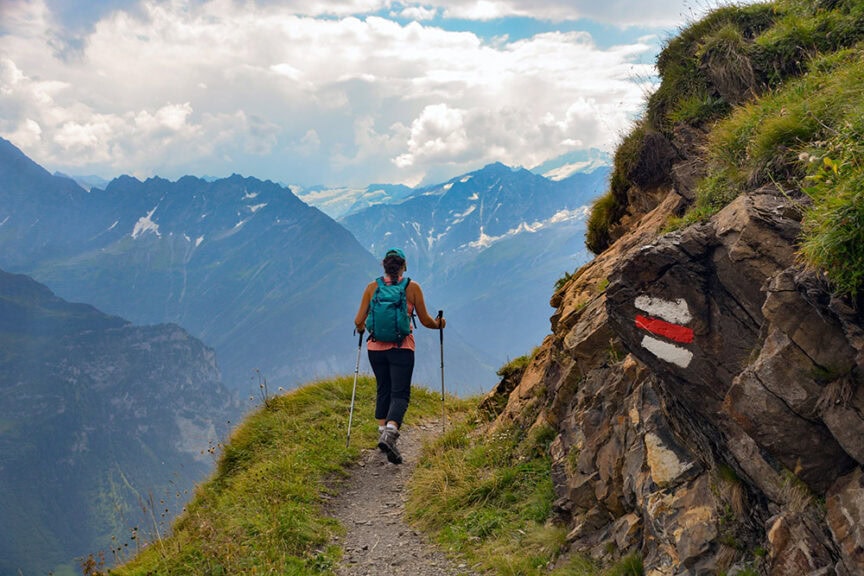 A person with hiking gear walks along a narrow mountain trail marked with a red and white trail sign, surrounded by the breathtaking scenery of the Swiss Alps, following the famous Via Alpina route.