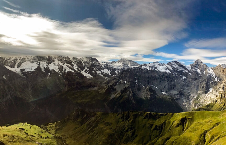 A panoramic view of Switzerland's mountain range with snow-capped peaks, green valleys, and a partly cloudy sky along the Via Alpina. The landscape features a mix of sunlight and shadows, perfect for hiking enthusiasts.