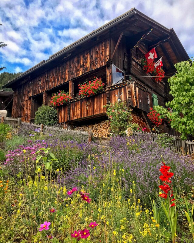 A rustic wooden house adorned with red flowers and a Swiss flag sits amidst a colorful flower garden under a partly cloudy sky, perfect for hikers exploring the Via Alpina in Switzerland.