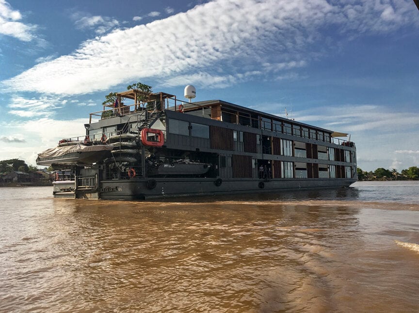 A large riverboat with multiple decks sails on the wide, muddy Mekong River in Cambodia under a partly cloudy sky, offering an unforgettable adventure.
