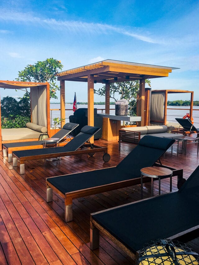 Outdoor lounge area with wooden deck, black sun loungers, and shaded daybeds. Overlooks the serene Mekong River with trees in the background under a sunny sky, offering a hint of adventure in Cambodia's natural beauty.