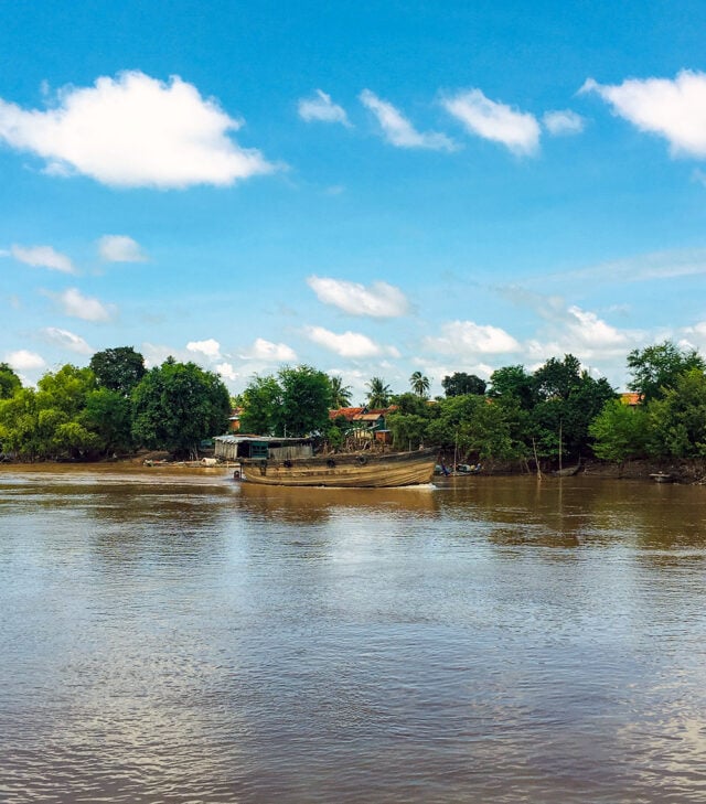A boat is docked by the shore of the Mekong River with trees and houses in the background under a blue sky with white clouds, inviting an adventure through Cambodia's scenic landscapes.