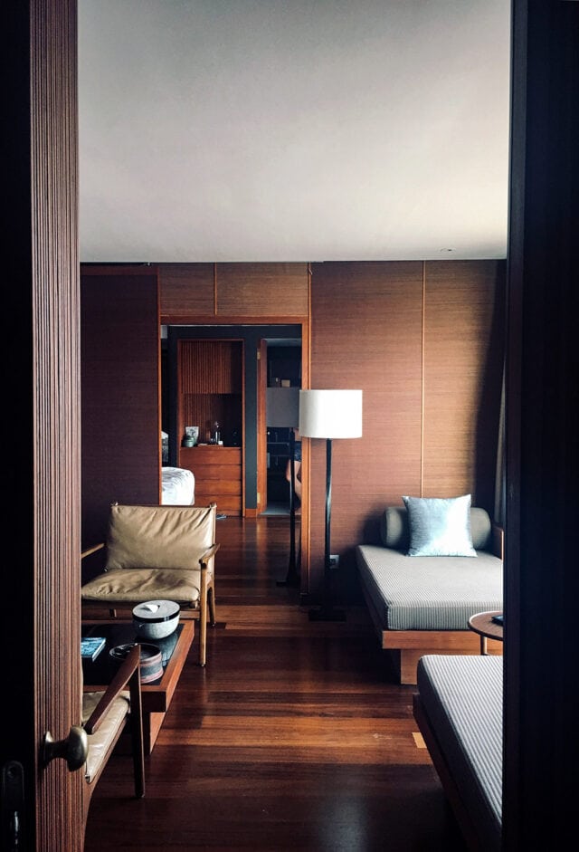 A room featuring wooden furniture, a floor lamp, two cushioned chairs with pillows, and decorative bowls on a small table. The room evokes the spirit of an adventure along the Mekong River and leads into another space visible through an open doorway.