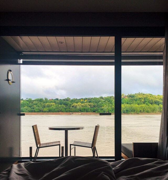 View of the Mekong River and green hills from a window with a balcony, featuring two chairs and a small round table in the foreground. This scenic snapshot perfectly captures the essence of adventure in Cambodia.