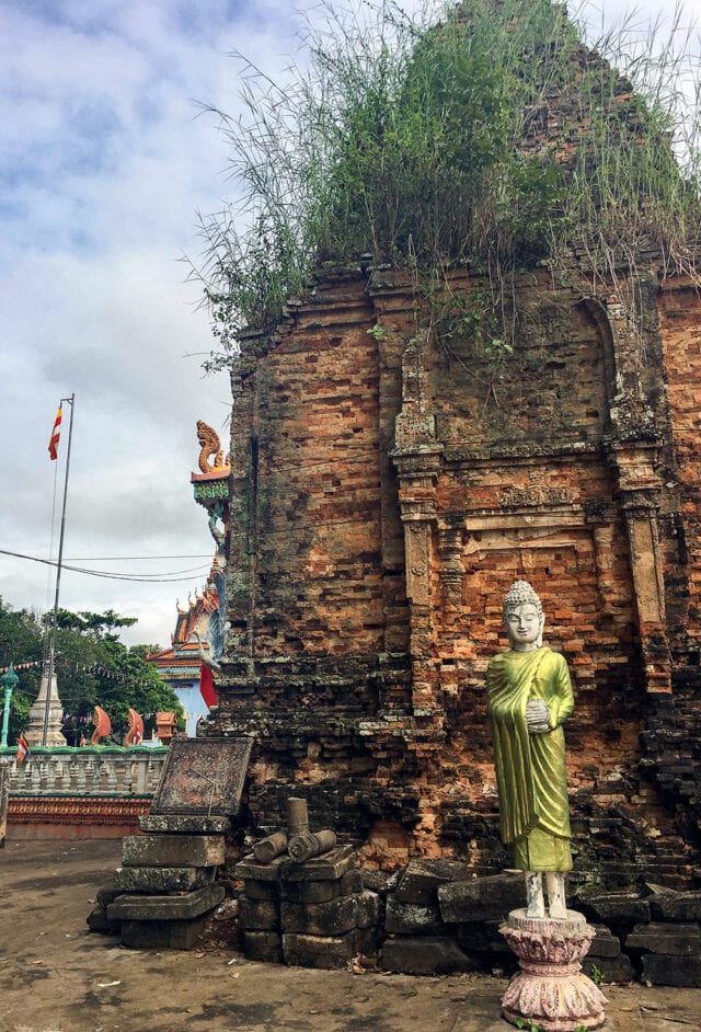 A small, aged brick shrine overgrown with grass houses a statue of a Buddha in a green robe standing on a pedestal, reminiscent of serene scenes by the Mekong River. Adjacent structures and a flagpole are visible in the background.