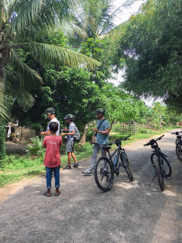 Three adults with bicycles and wearing helmets stand on a road surrounded by lush greenery in Cambodia, while a young boy in a red shirt looks on. One adult appears to be pointing at something off-camera, perhaps an exciting spot along the Mekong River—perfect for their next adventure.