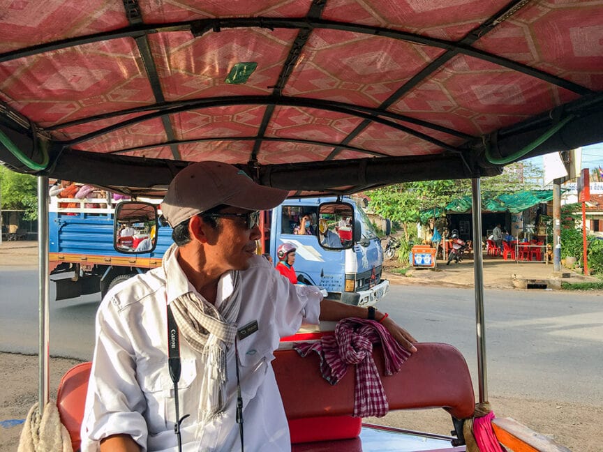A man wearing a white shirt and cap, with a camera strap around his neck, is seated under a canopy in a vehicle, looking to his left. It appears he's capturing moments of an adventure along the Mekong River in Cambodia. In the background, people and vehicles are visible on the road.