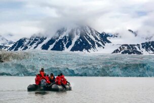 A group of people in red jackets sit on an inflatable boat on a body of water with a mountainous, snow-covered landscape and glacier in the background. This Svalbard wildlife adventure showcases the stunning beauty of the polar region.