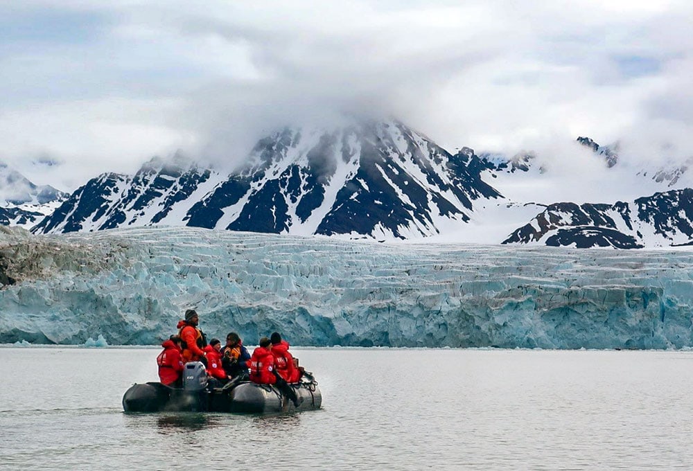 A group of people in red jackets sit on an inflatable boat on a body of water with a mountainous, snow-covered landscape and glacier in the background. This Svalbard wildlife adventure showcases the stunning beauty of the polar region.
