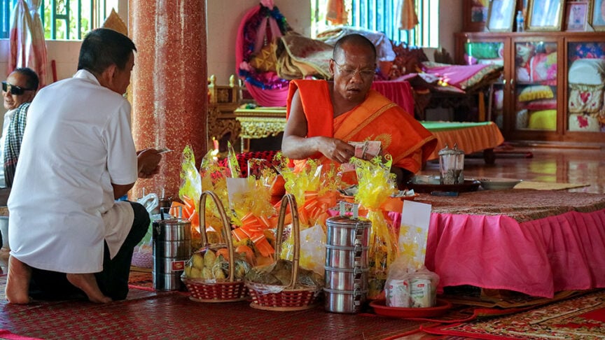 A monk in orange robes and a man in white sit on the floor of a temple in Cambodia, engaged in activities involving food offerings and wrapped items. A pink cloth covers a table in the background, adding to the serene atmosphere of this spiritual adventure near the Mekong River.