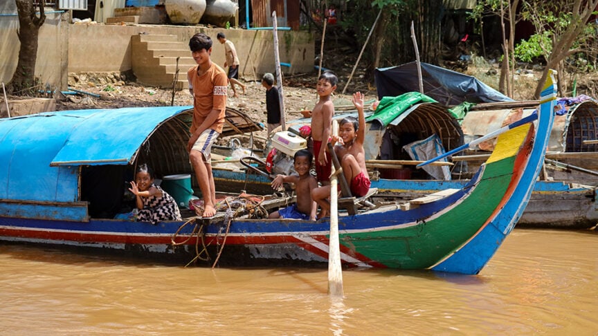 Children on a colorful wooden boat, some sitting and some standing, embark on an adventure along the muddy Mekong River with the small huts of Cambodia in the background.