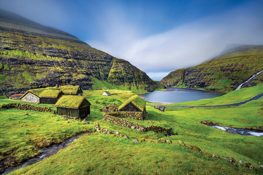 Small grass-roofed houses nestled in a green valley by a lake, surrounded by tall, rocky hills under a cloudy sky—a tranquil contrast to the vibrant energy of colorful cities often featured in world travel.